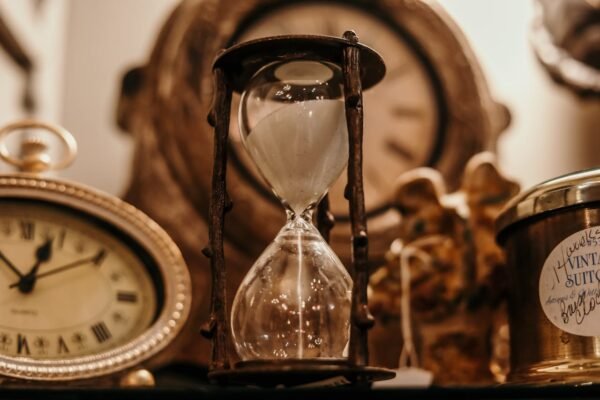 Close-up of a vintage hourglass surrounded by antique clocks in warm lighting.